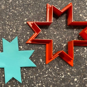 May include: Two star-shaped cookie cutters, one red and one light blue. The red cutter is larger and has a more intricate design.