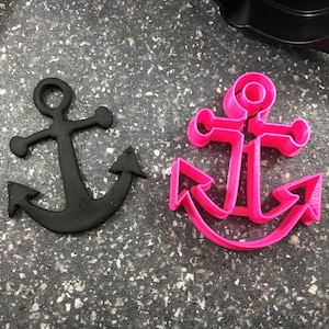 May include: Two anchor-shaped cookie cutters, one black and one pink. The black cutter is solid, while the pink cutter is hollow.