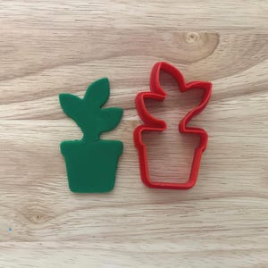 May include: A green plant-shaped cookie cutter and a red plant-shaped cookie cutter sit on a wooden surface. The green plant is a solid shape, while the red one is an outline.