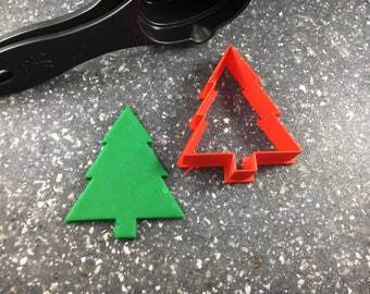 May include: Two Christmas tree cookie cutters, one green and one red. The green cutter is solid, while the red cutter is hollow.