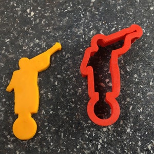 May include: Two trumpet-shaped cookie cutters. One is solid orange, and the other is red with a hollow center. The cutters are designed in the shape of a person playing a trumpet, with a circular base.