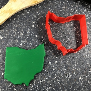 May include: A green Ohio state cookie cutter and a red Ohio state cookie cutter on a gray countertop.