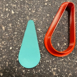 May include: Two cookie cutters, one shaped like a teardrop and the other shaped like a rounded oval. The teardrop cutter is light blue and the oval cutter is red.
