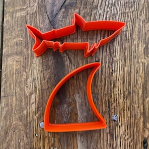 May include: Two orange plastic cookie cutters shaped like a shark and a shark fin.