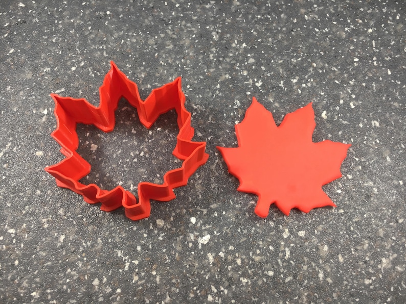 May include: Two red plastic maple leaf cookie cutters. One is a solid leaf shape and the other is an outline of a leaf.