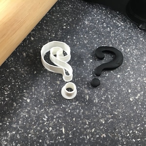 May include: Two question mark cookie cutters, one white plastic and one black plastic, on a gray speckled surface.