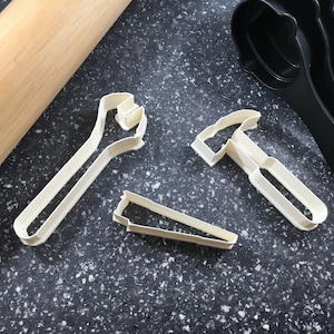 Puede incluir: Tres cortadores de galletas de plástico blanco con forma de llave inglesa, martillo y sierra. Los cortadores están sobre una superficie gris moteada.