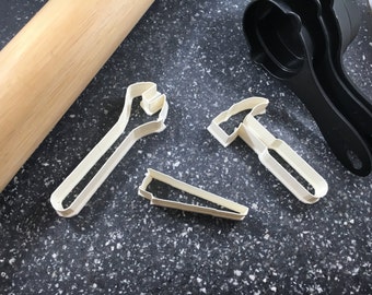 May include: Three white plastic cookie cutters shaped like a wrench, a hammer, and a saw. The cutters are on a gray speckled surface.