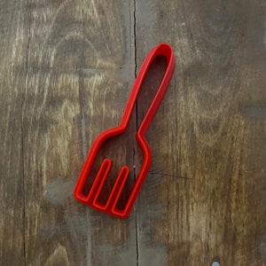 May include: Red plastic fork cookie cutter. The cutter is shaped like a fork with three tines.