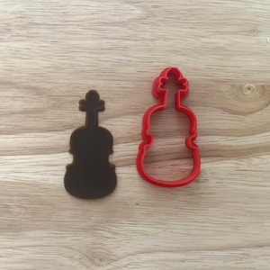 May include: Two violin-shaped objects on a wooden surface. One is a solid dark brown silhouette, and the other is a red outline of a violin. The objects are likely cookie cutters or craft supplies.