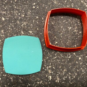 May include: An orange, square-shaped cookie cutter with rounded corners sits next to a teal-colored, square-shaped object with rounded corners. The cookie cutter is made of plastic and has a textured surface.