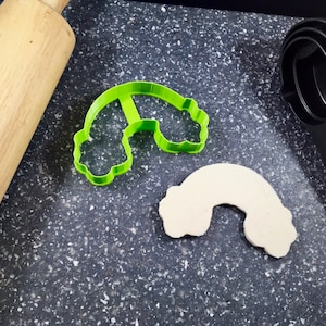 May include: A green plastic rainbow cookie cutter and a white fondant rainbow cookie on a black speckled surface. A wooden rolling pin and a black measuring cup are also visible.