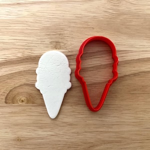 Könnte beinhalten: Ein roter Eistüten-Ausstecher aus Kunststoff neben einer weißen Eisform auf einer Holzoberfläche. Der Ausstecher ist eine detaillierte Umrisszeichnung einer Eistüte.