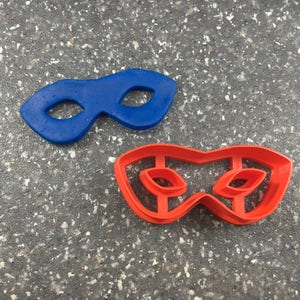 May include: Two plastic cookie cutters shaped like superhero masks. One is blue and the other is red.