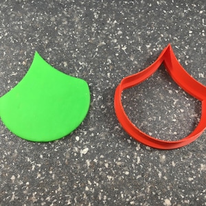 May include: A green plastic cookie cutter shaped like a teardrop, next to a red plastic cookie cutter shaped like a teardrop.