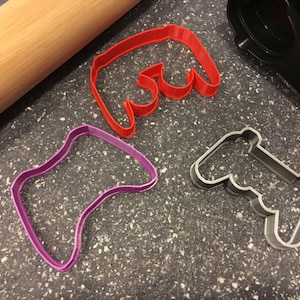 Puede incluir: Tres cortadores de galletas diferentes con forma de mandos de videojuegos. Los cortadores son rojo, morado y gris.