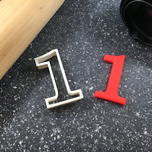 May include: Two number one cookie cutters, one white and one red, on a black speckled surface. A wooden rolling pin is in the background.