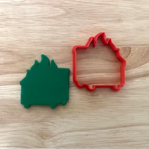 May include: A green and red cookie cutter set in the shape of a house on fire. The green cookie is a solid shape, while the red one is an outline. Both are on a wooden surface.
