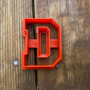May include: A red plastic cookie cutter shaped like the letter D. The cookie cutter is on a wooden surface.