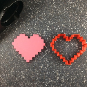 May include: A pink pixelated heart and a red heart-shaped cookie cutter sit on a speckled gray surface. The cookie cutter is made of red plastic and has a pixelated design. The pink heart is a solid, pixelated shape.