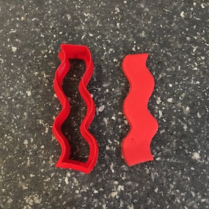 Puede incluir: Dos cortadores de galletas de plástico rojo en forma de líneas onduladas. El cortador de la izquierda es una forma cerrada, mientras que el cortador de la derecha es una forma abierta.