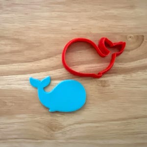 May include: Two whale-shaped cookie cutters. One is a solid, bright blue whale. The other is a red outline of a whale. Both are on a light brown wooden surface.