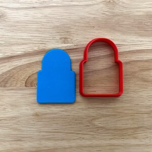 May include: A blue, rectangular cookie cutter with a rounded top, next to a red cookie cutter in the same shape. The cutters are on a light brown wooden surface.