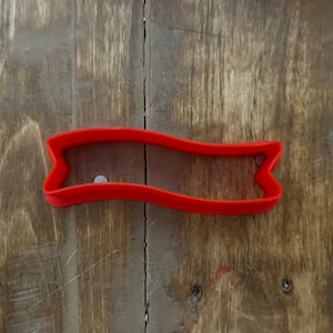 Może przedstawiać: Ciastko wykrawacz z czerwonego plastiku w kształcie banera z falistym górnym i dolnym brzegiem.