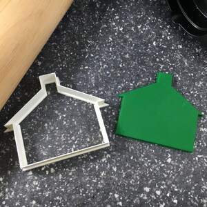 Puede incluir: Un cortador de galletas de plástico blanco con forma de casa, junto a un cortador de galletas de plástico verde con forma de casa.