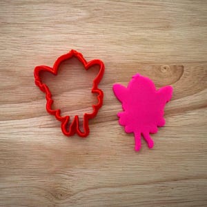 May include: A red plastic cookie cutter in the shape of a fairy, next to a pink fairy-shaped cookie. The cutter has a detailed outline, and the cookie is a solid color. Both are on a wooden surface.