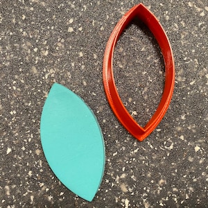 May include: A teal-colored, leaf-shaped object next to a red, leaf-shaped cookie cutter. The cookie cutter is made of plastic and has a smooth, rounded shape. The teal object appears to be made of wood or clay.