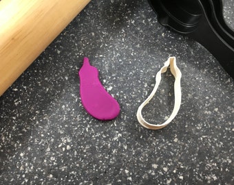 May include: A purple clay eggplant shape next to a white plastic eggplant cookie cutter on a black speckled surface.