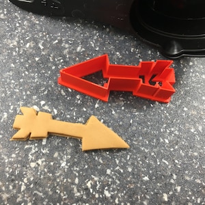 Puede incluir: Un cortador de galletas de flecha de plástico rojo y una galleta de flecha marrón sobre una superficie gris moteada.