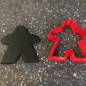 Puede incluir: Una pieza de juego negra y un cortador de galletas rojo con forma de persona. La pieza de juego es de color negro sólido, mientras que el cortador es de plástico rojo. Los artículos están sobre una superficie gris moteada. Los artículos están relacionados con los juegos de mesa.