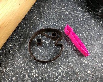 May include: A brown cookie cutter in the shape of a painter's palette with two circular wells and a pink paintbrush-shaped tool. The items are on a speckled gray surface.