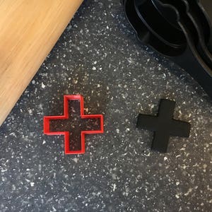 May include: Two plastic cookie cutters in the shape of a plus sign. One is red and the other is black.