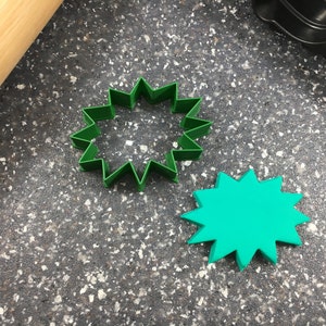May include: Two green star-shaped cookie cutters on a gray speckled surface. The cutter on the left is open and the cutter on the right is closed.