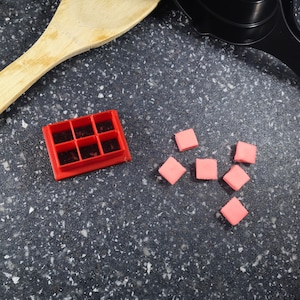May include: A red plastic mold with six square compartments, and six pink square-shaped candies on a black speckled surface.