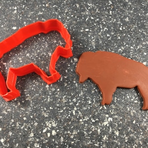 Puede incluir: Cortador de galletas de plástico rojo con forma de bisonte, junto a un recorte de bisonte de arcilla marrón.