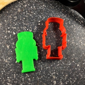May include: Two plastic cookie cutters in the shape of a soldier. One is bright green, and the other is red. The cutters are on a speckled gray surface, with a wooden spoon and a black spatula in the background.