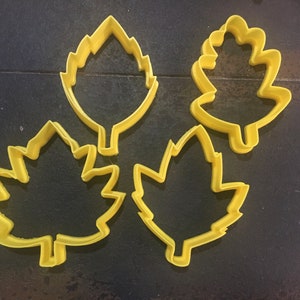 Könnte beinhalten: Vier gelbe, plastikförmige Blätter-Ausstecher. Die Ausstecher sind in Form eines Ahornblattes, eines Eichenblattes und zwei verschiedenen Arten von Blättern.