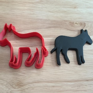May include: A red donkey-shaped cookie cutter and a black donkey-shaped object are displayed on a wooden surface. The cookie cutter is made of plastic and the donkey object appears to be made of a solid material.