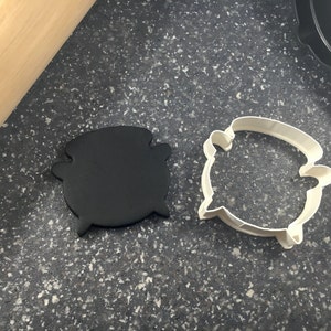 Könnte beinhalten: Ein schwarzer Kessel-Ausstecher und ein schwarzer Fondant-Kessel auf einer grauen, gesprenkelten Oberfläche.