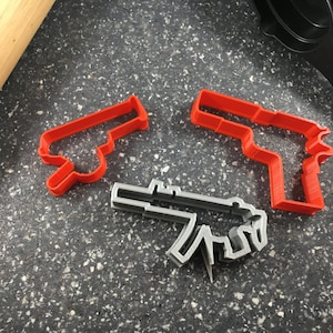 May include: Three cookie cutters in the shape of firearms. Two are red, depicting handguns, and one is grey, shaped like a submachine gun. The cutters are on a speckled grey surface, near a wooden rolling pin.