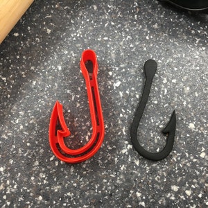 May include: Two fishing hook shaped cookie cutters, one red and one black, on a gray speckled surface.