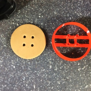 May include: A close-up of a round orange cookie cutter with four holes in the center. Next to it is a round orange cookie with four holes in the center.
