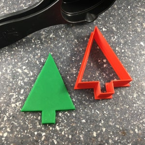 May include: A green Christmas tree-shaped cookie cutter next to a red tree-shaped cookie cutter. A black measuring spoon is in the background. The cookie cutters are on a speckled gray surface.