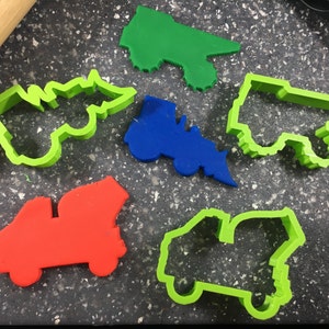May include: Five different cookie cutters shaped like dump trucks. The cutters are made of green, blue, and red plastic.