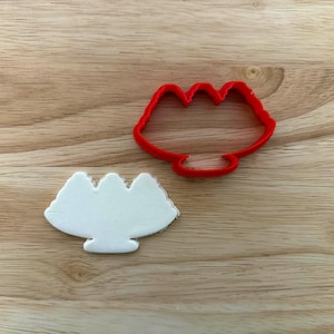 May include: A red plastic cookie cutter in the shape of a mouth, next to a white cookie in the same shape. The cookie cutter has a curved bottom and three peaks at the top. The cookie has a similar shape.