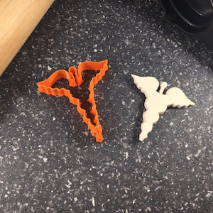May include: Two plastic cookie cutters in the shape of the medical symbol, one orange and one white, on a dark grey speckled surface.